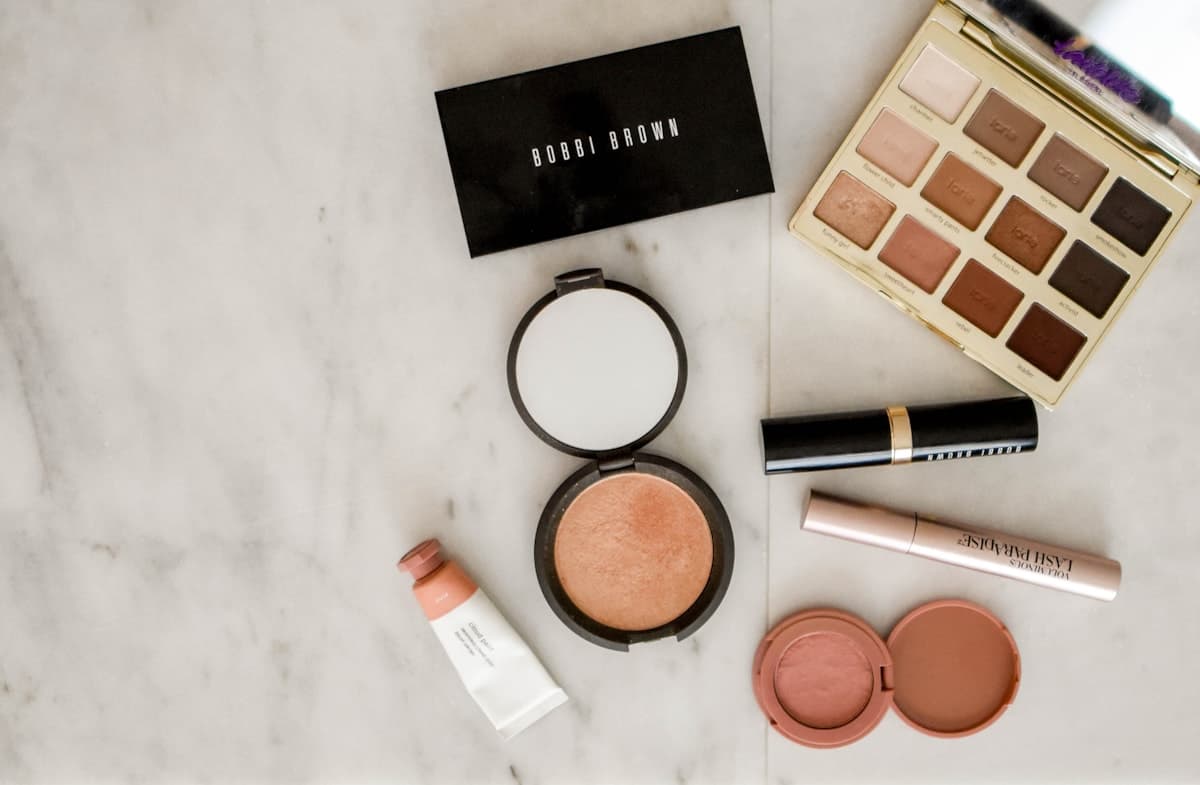 Makeup on a marble surface, including an eyeshadow palette, a bronzer compact, mascara and a pink cream blush